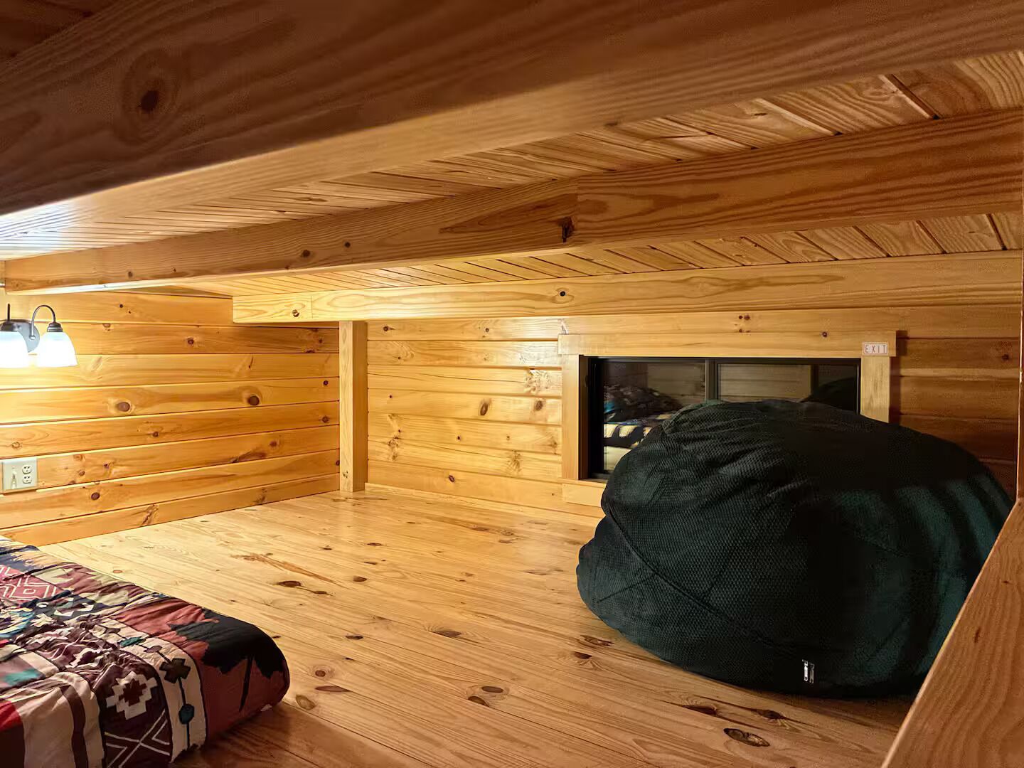 Une pièce chaleureuse dans un chalet, avec un plancher en bois, un lit recouvert d’une couverture à motifs, un fauteuil en forme de poire et une cheminée.