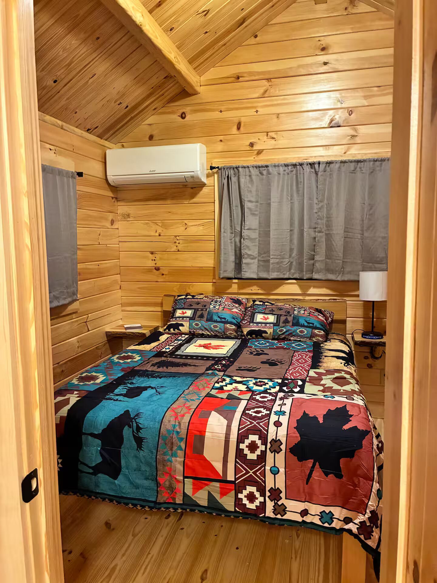 Una habitación de cabaña acogedora con interior de madera, una cama con colcha colorida, una ventana con cortinas y un aire acondicionado de pared.