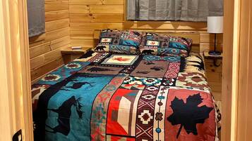 Una habitación de cabaña acogedora con interior de madera, una cama con colcha colorida, una ventana con cortinas y un aire acondicionado de pared.