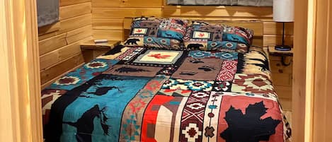 Una habitación de cabaña acogedora con interior de madera, una cama con colcha colorida, una ventana con cortinas y un aire acondicionado de pared.