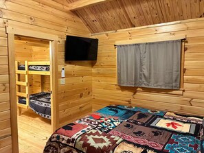 Une chambre de style cabane en bois, avec un lit superposé, un téléviseur à écran plat, une fenêtre avec un rideau et une couverture colorée sur le lit.