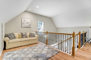 En loftstue med højt til loftet, en trappe i træ med sorte spær, en beige sofa med mønstrede puder og et stort vindue med persienner.
