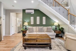 A living room with a sofa, coffee table, and decorative wall art. A staircase with a railing is visible in the background.