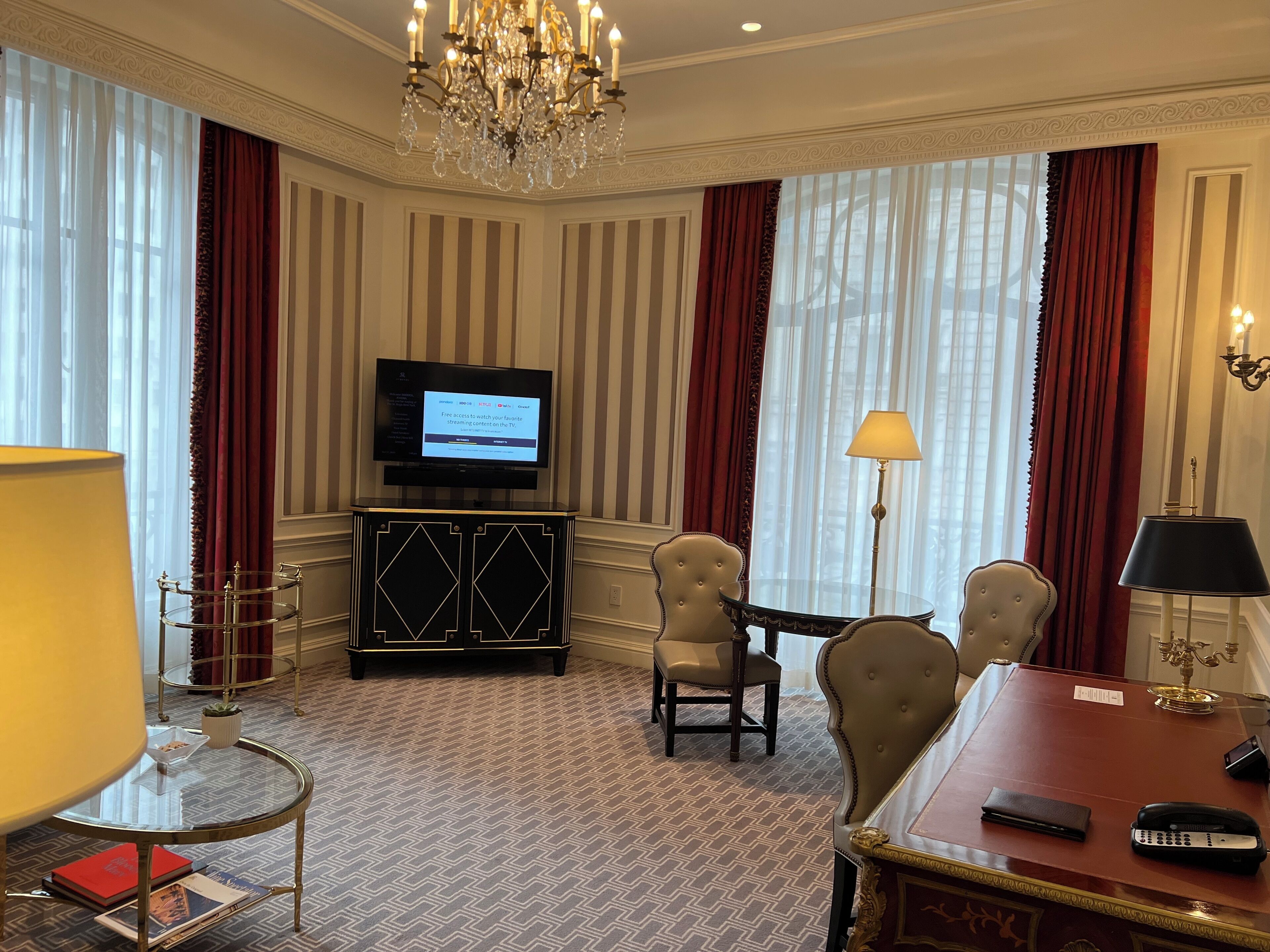 A hotel room with a television, two armchairs, a desk with a telephone, and a chandelier.