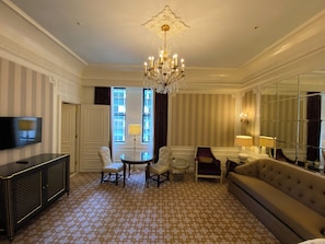 Quarto de hotel com lustre, sofá, televisão e área de jantar com cadeiras e mesa.