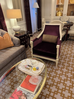 Uma sala de estar com um sofá estampado, uma poltrona roxa, uma mesa de centro de vidro com um tabuleiro de objetos e um tapete estampado.