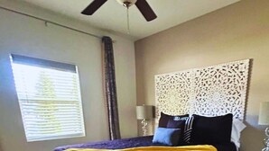 Quarto com cama, mesa de cabeceira, persianas na janela e cabeceira decorativa.