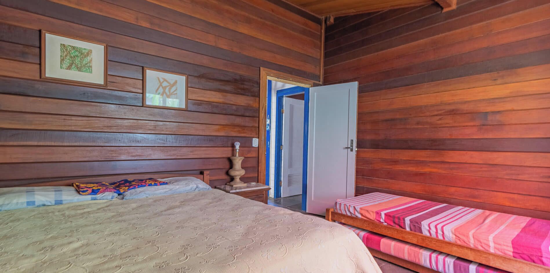 Una habitación con paredes de madera, una cama con colcha estampada, una mesita de noche con lámpara y una puerta que da a otra habitación.