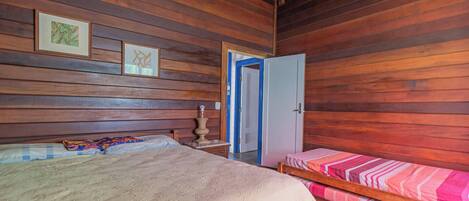 Una habitación con paredes de madera, una cama con colcha estampada, una mesita de noche con lámpara y una puerta que da a otra habitación.