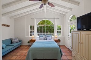 Un dormitorio con cama, un sofá azul, televisor y ventana con vista a follaje.
