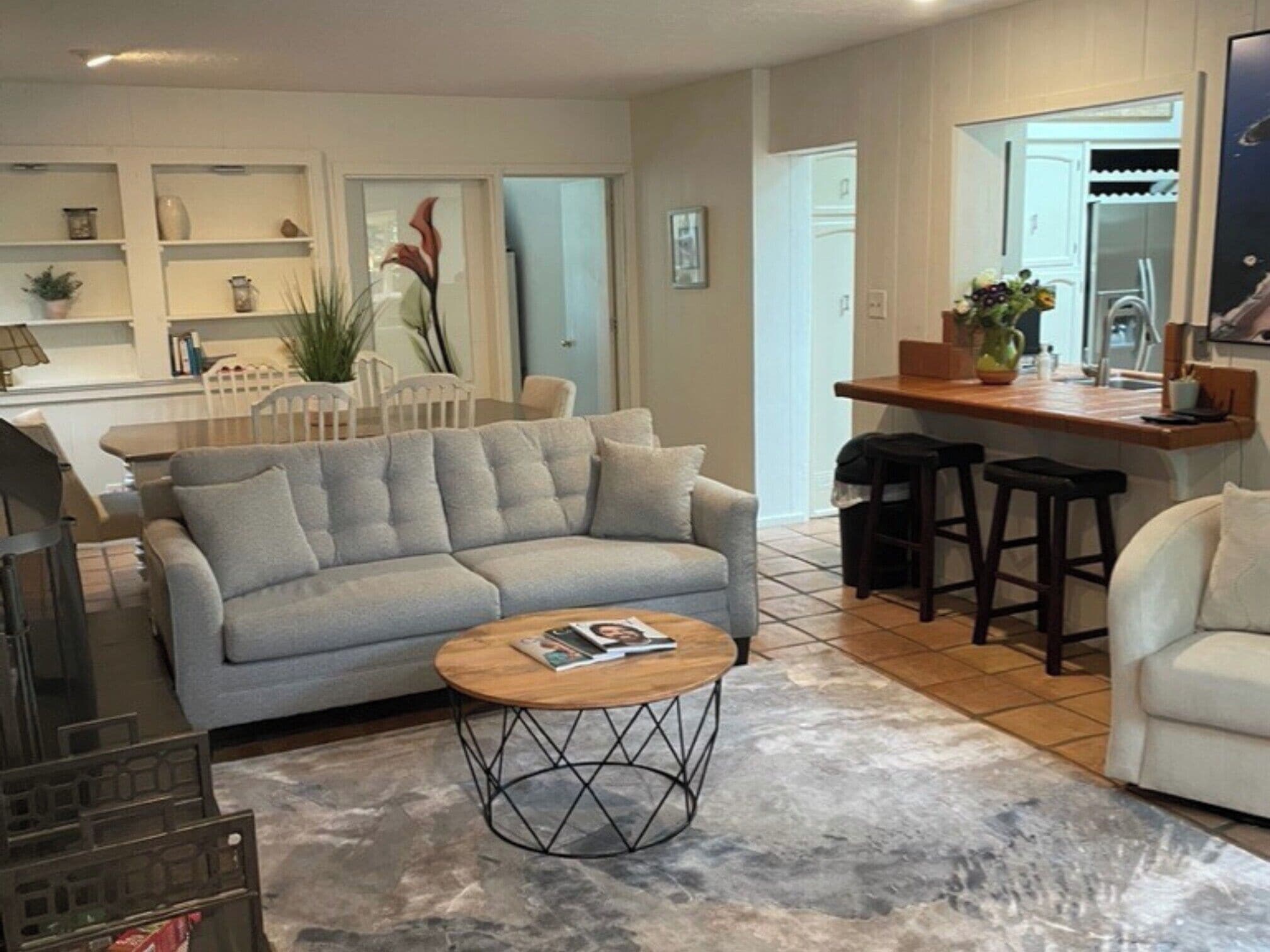 Ein Wohnzimmer mit grauem Sofa, einem Couchtisch und einer Küchenzeile mit Arbeitsfläche und Hockern.