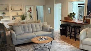 Una sala de estar con un sofá gris, una mesa de centro y una cocina con mostrador y taburetes.