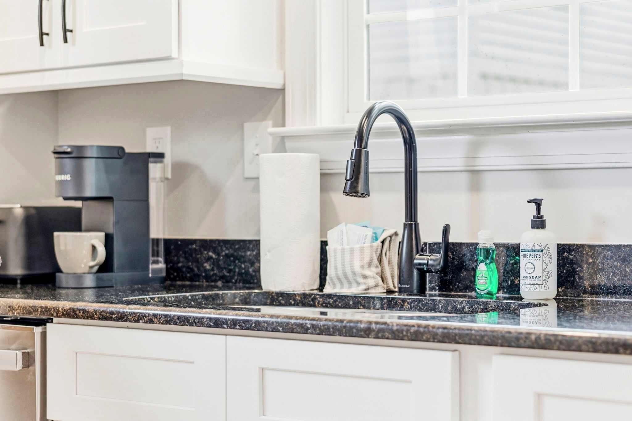 A kitchen with a black countertop, white cabinets, a coffee maker, a paper towel holder, a soap dispenser, and a window.