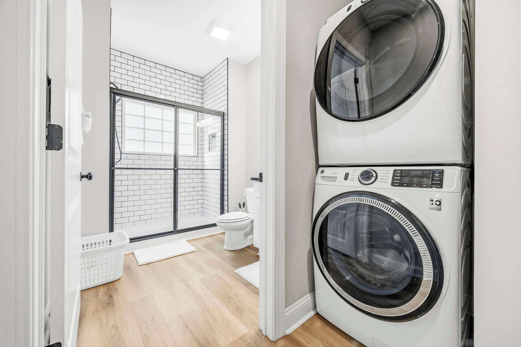 A laundry room with two stacked front-loading washing machines, a white toilet, and a window with a view of the outside.