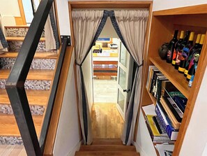 Uma escada com corrimão de madeira, uma estante com diversos objetos e uma porta que dá para outro quarto.