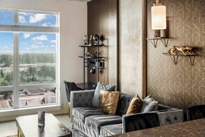 A modern living room with a sofa, a coffee table, and a view of a cityscape through large windows. - Terra - Luxe 1BR Condo w/ Sunset Views, Pool, Gym (Orlando)