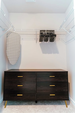 A walk-in closet with a dark wooden dresser, a patterned wall hanging, and a metal wire shelf with hanging items. - Terra - Luxe 1BR Condo w/ Sunset Views, Pool, Gym (Orlando)