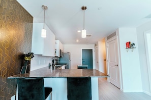 A modern kitchen with a central island, pendant lighting, and a patterned wall. - Terra - Luxe 1BR Condo w/ Sunset Views, Pool, Gym (Orlando)