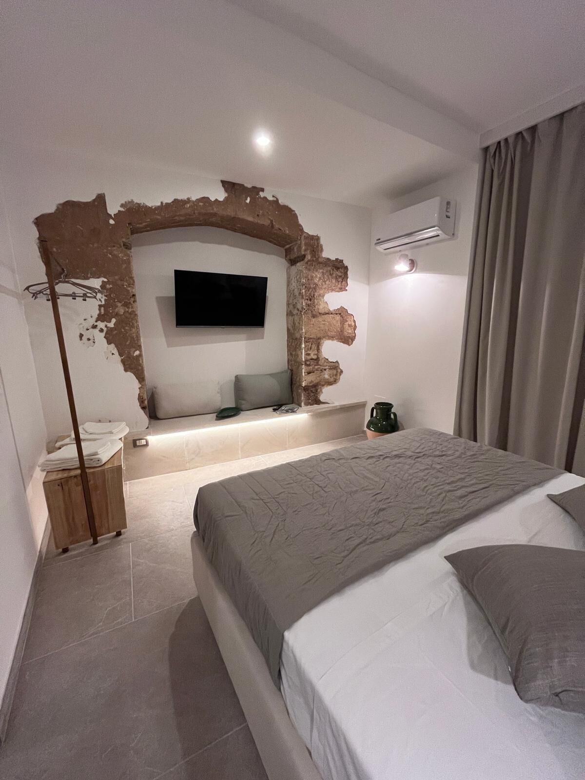 Une chambre à coucher avec un grand lit, un téléviseur à écran plat fixé à un mur en briques, une table de chevet et une lampe sur pied.