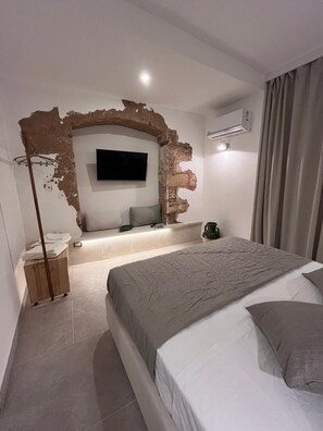 A bedroom with a large bed, a flat-screen TV mounted on a brick wall, a bedside table, and a floor lamp. - Vrbo Property (Marina di Mancaversa)
