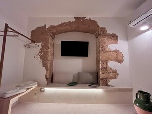 A bathroom with a large, unfinished wall arch, a flat-screen TV mounted above a bench, and a green towel on the bench. - Ada Vacation Home • 2 minutes from the ocean • Relaxation for Couples (Marina di Mancaversa)