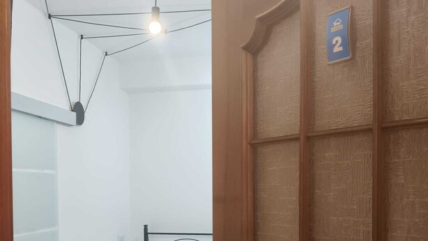Quarto com uma cama, porta com placa de número e luminária pendente.