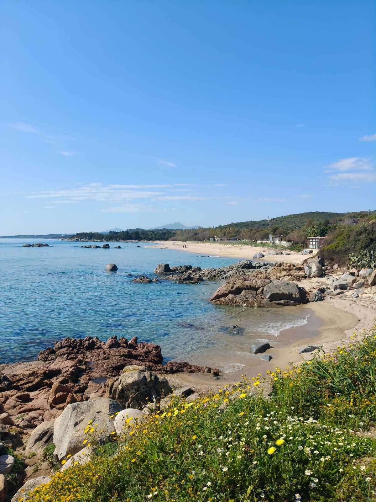 Paisatge costaner amb cels clars, línia de costa rocosa i platja sorrenca.