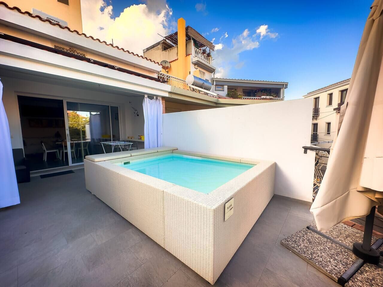 Ein rechteckiger Pool mit weißem Rand, umgeben von einer überdachten Terrasse mit Tisch und Stühlen.