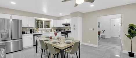 Une cuisine moderne avec des meubles blancs, une table à manger pour quatre personnes et un ventilateur de plafond.