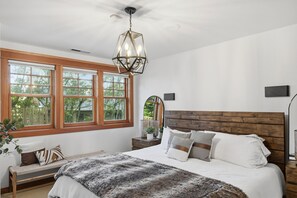 Ein Schlafzimmer mit einem großen Bett, einem hölzernen Kopfteil, einer Bank mit Kissen, einer Pendelleuchte und einem Fenster mit zahlreichen Sprossen.