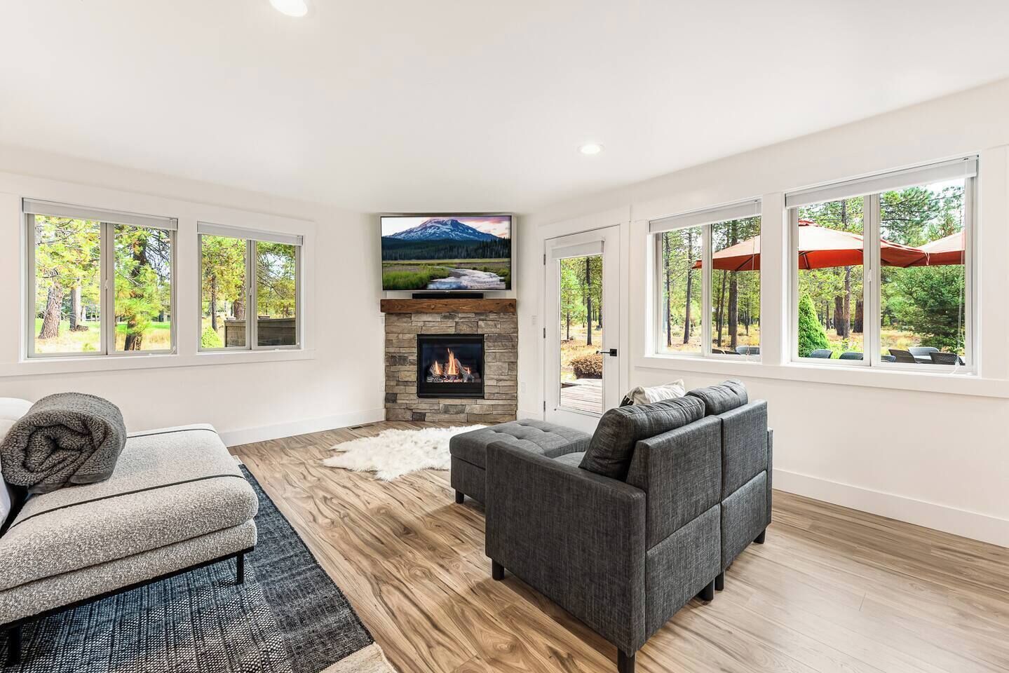 A modern living room with a fireplace, a grey sofa, and a large window overlooking a forest.