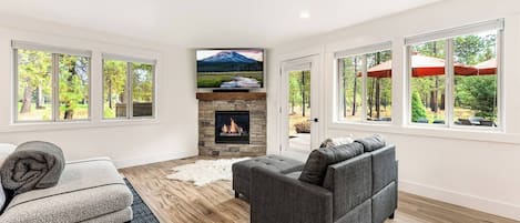 A modern living room with a fireplace, a grey sofa, and a large window overlooking a forest.
