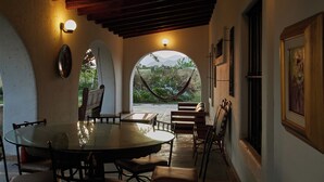 Un porche techado con mesa y sillas, una hamaca, y vistas a la vegetación y montañas.