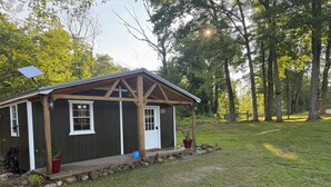 A small wooden structure with a porch, a door, and windows, surrounded by a grassy area and trees.
