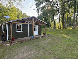A small wooden structure with a porch, a door, and windows, surrounded by a grassy area and trees.