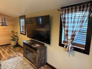 A living room with a flat-screen TV mounted on the wall, a wooden cabinet with a TV stand, a potted plant, and a window with checkered curtains.