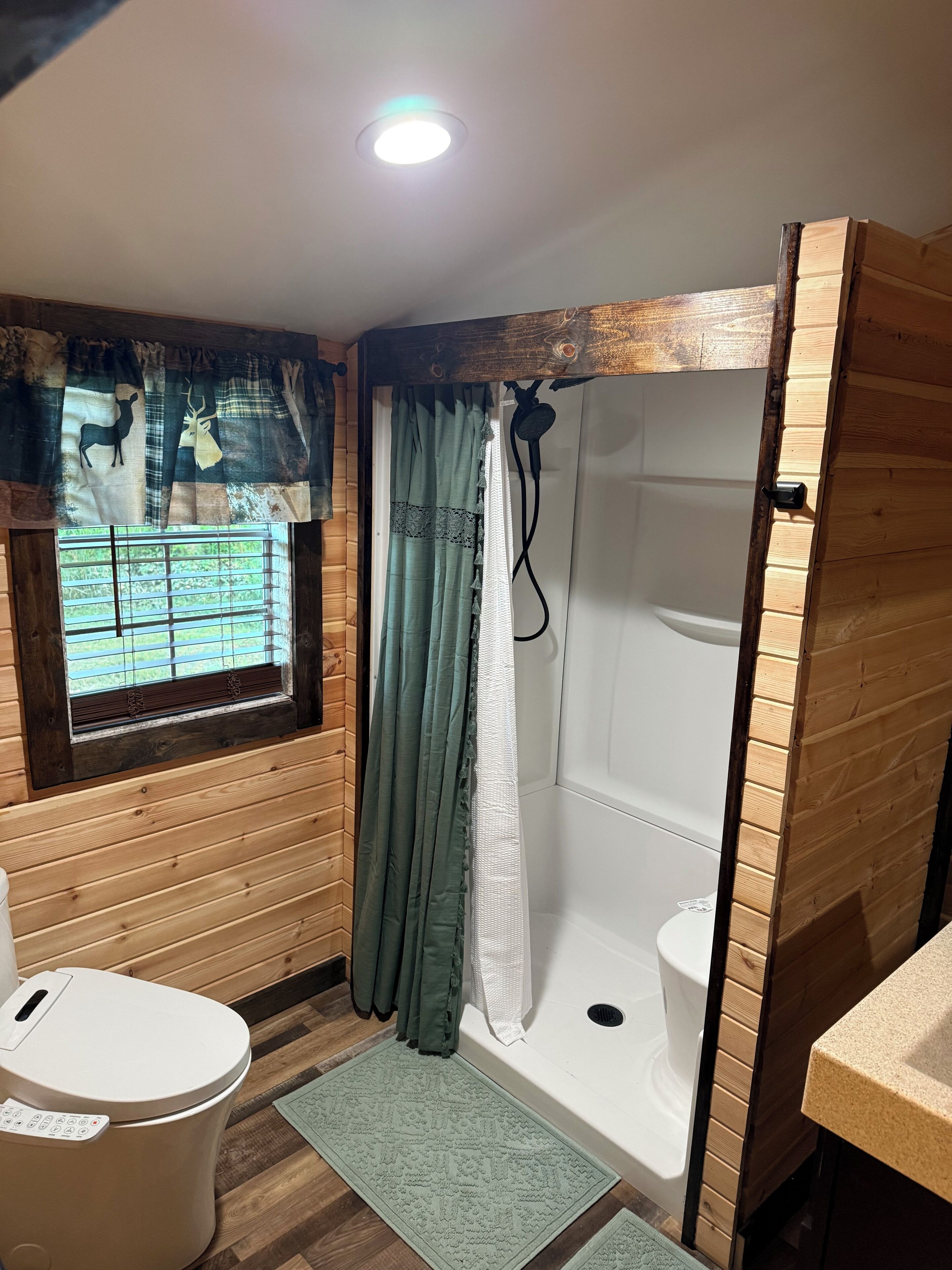 A bathroom with a wooden interior, a window with blinds, a shower, and a toilet.