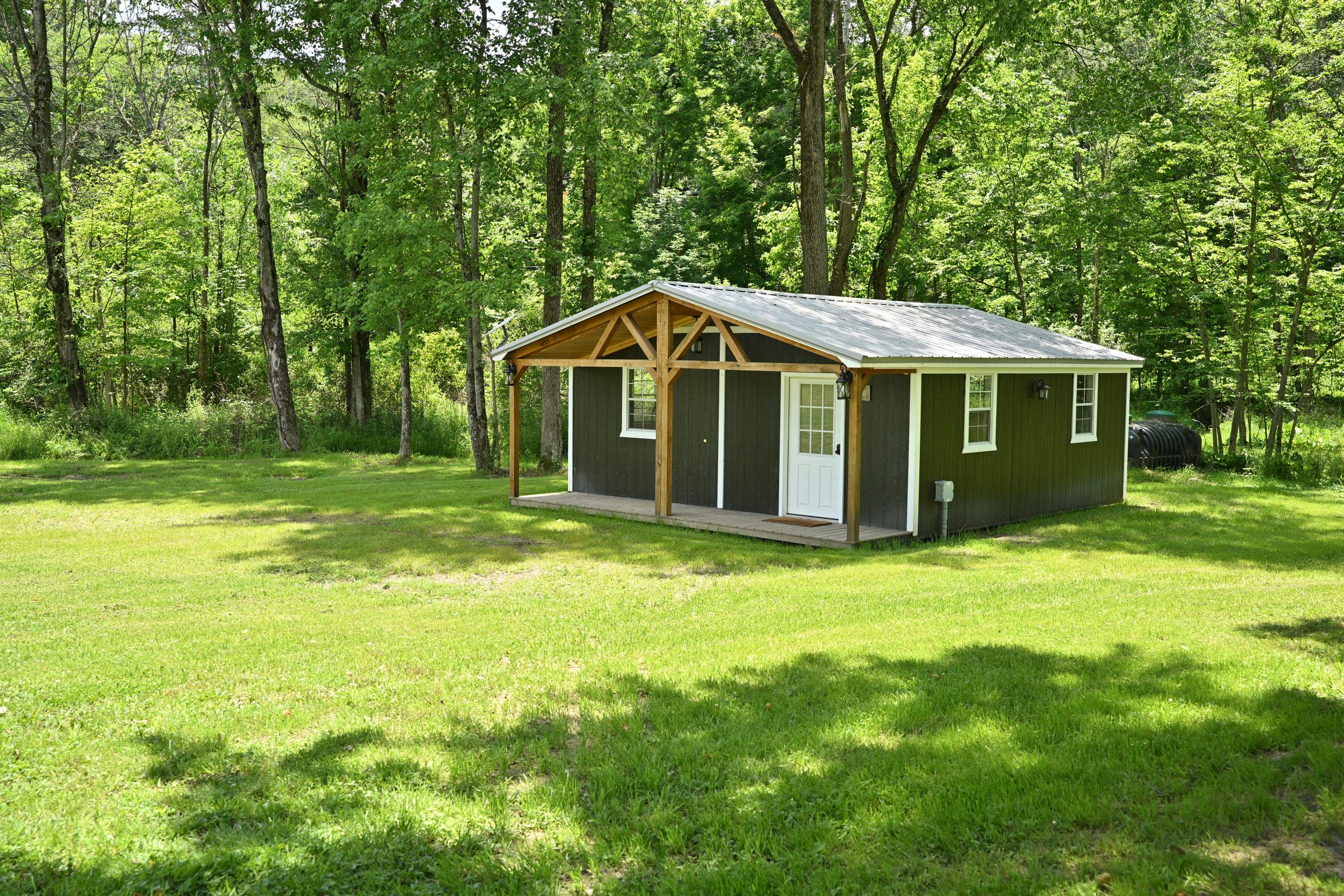 A small wooden cabin with a porch, surrounded by a grassy field and dense trees.