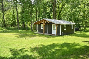 A small wooden cabin with a porch, surrounded by a grassy field and dense trees.
