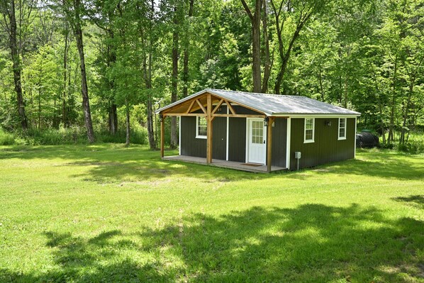 A small wooden cabin with a porch, surrounded by a grassy field and dense trees.