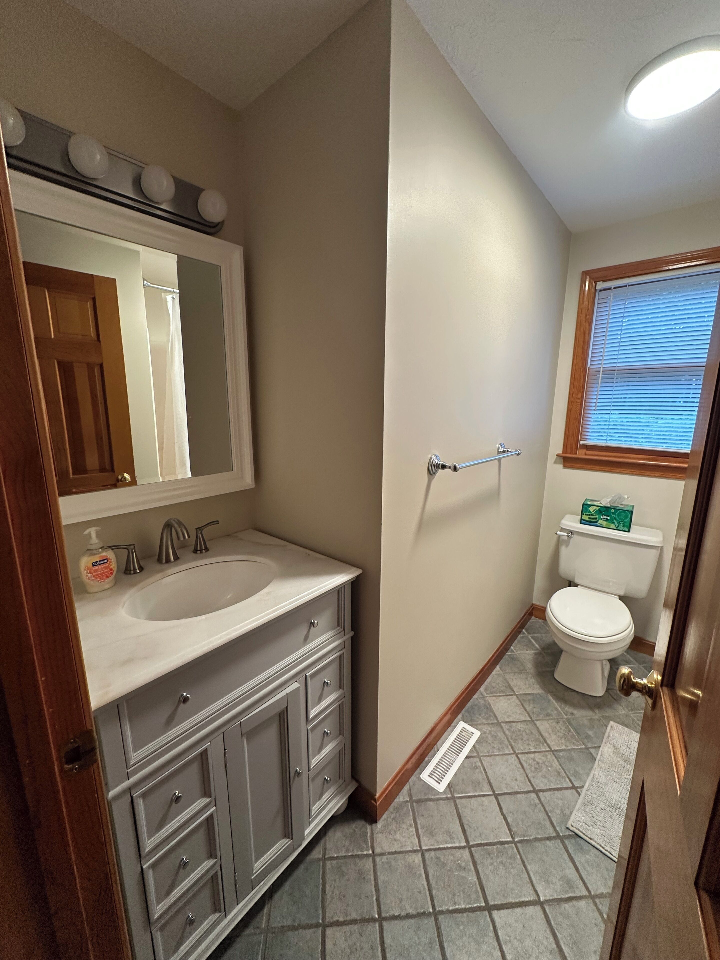 A bathroom with a double sink vanity, a toilet, a window with blinds, and a shower area.