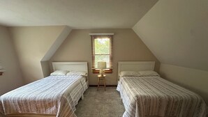 Quarto com duas camas, janela com persianas, uma mesa pequena com abajur e um tapete estampado.