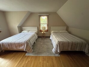 Un dormitorio con dos camas, una ventana con persianas, una mesita con lámpara y una alfombra estampada.