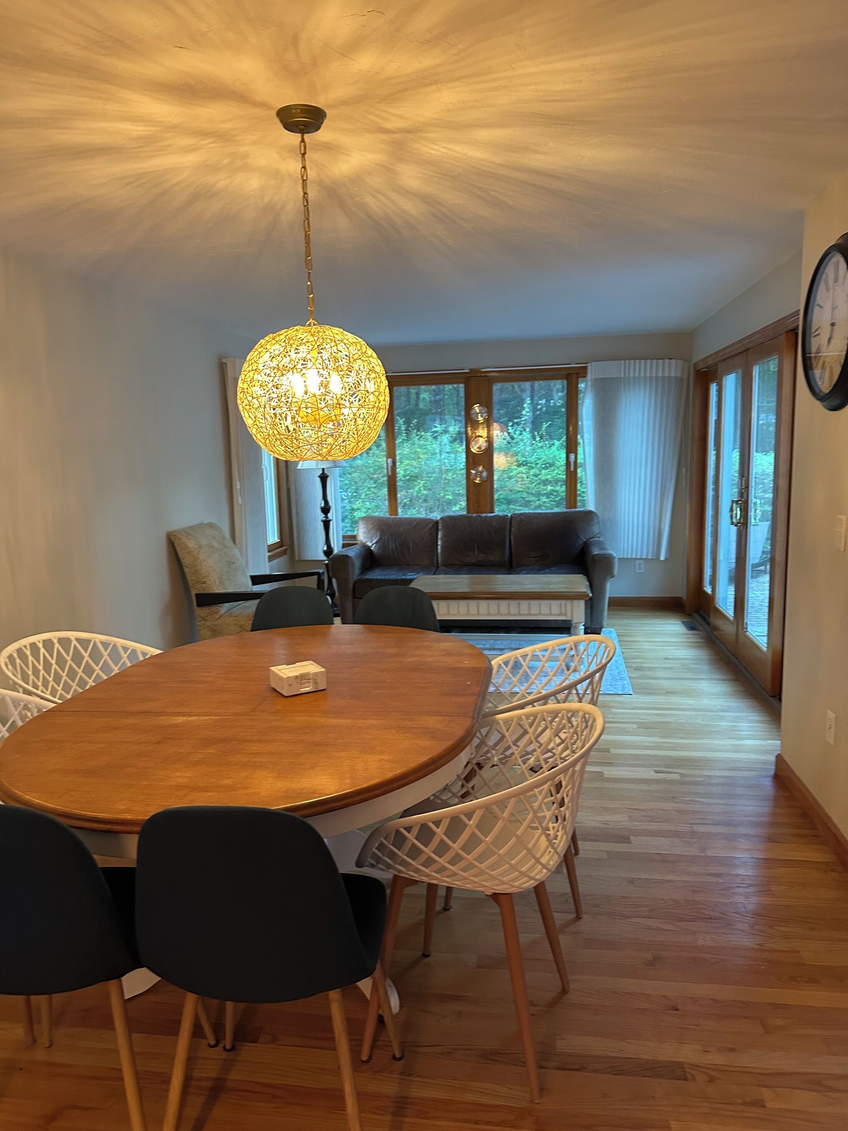 A dining area with a round wooden table, white wicker chairs, a hanging light fixture, and a view of the outdoors through large windows.