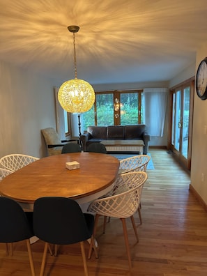 Un coin repas avec une table ronde en bois, des chaises en osier blanches, un luminaire suspendu et une vue sur l’extérieur grâce à de grandes fenêtres.