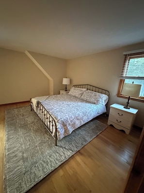A bedroom with a bed, nightstands, a lamp, a window, and a wooden floor. - Heart of the Bay! Charming 3-bedroom house with AC, walk to the beach. (Mashpee)