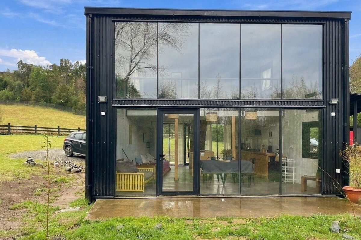 Un edificio moderno con estructura de vidrio y marcos negros, donde se distingue el interior con un comedor y una zona de estar.