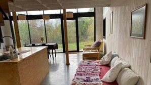 A room with a wooden dining table, a sofa with cushions, and a view through large windows.