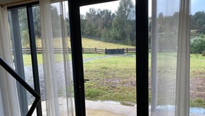 A view through a glass door with a metal frame, leading to an outdoor area with a grassy field, a wooden fence, and a paved path.