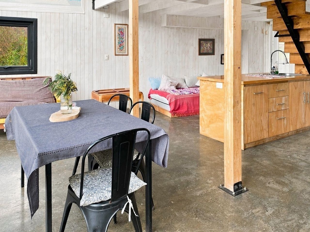 A dining area with a table set for two, a wooden bench, and a kitchen area with a bed in the background.